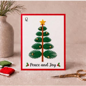 Peut inclure: Une carte de vœux blanche à bordure rouge ornée d'un sapin de Noël brodé en fil vert avec une étoile jaune, des perles dorées et un tronc marron. Le texte Peace and Joy est imprimé en noir avec de petites illustrations de houx.