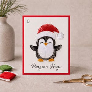 Op de afbeelding: Een witte wenskaart met een rode rand voorzien van een draadkunstontwerp van een pinguïn met een rood-witte kerstmuts. De pinguïn is zwart, wit en geel. De tekst "Penguin Hugs" staat in het zwart onder de pinguïn, met een kleine "Q" in de linkerbovenhoek.