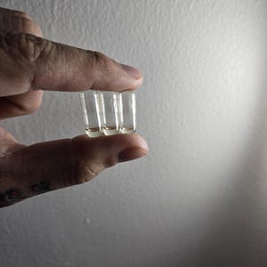 May include: Three small, clear glass vials are held between fingers against a neutral background. Each vial has a rounded bottom and a narrow opening. The vials appear to contain a clear liquid. The image is well-lit, highlighting the transparency of the glass.