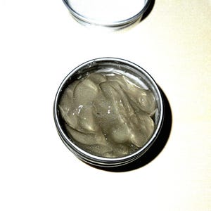 Könnte beinhalten: Ein kleines, offenes Silberdöschen gefüllt mit einem durchscheinenden, graugrünen Gel. Das Gel hat ein leicht strukturiertes Aussehen. Ein leerer Silberdöschen-Deckel liegt über dem offenen Döschen auf einer weißen Oberfläche. Das Produkt ist wahrscheinlich ein Hautpflegeartikel.