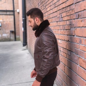 Puede incluir: Chaqueta bomber de cuero marr&oacute;n con cuello de piel marr&oacute;n oscuro. La chaqueta tiene dobladillo y pu&ntilde;os de canal&eacute;, dos bolsillos delanteros y cierre de bot&oacute;n. La persona est&aacute; de pie junto a una pared de ladrillos.