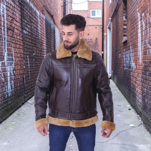 May include: A brown leather jacket with a tan shearling collar and cuffs. The jacket features a front zipper, a belt at the waist, and a classic aviator style. The model is wearing blue jeans.