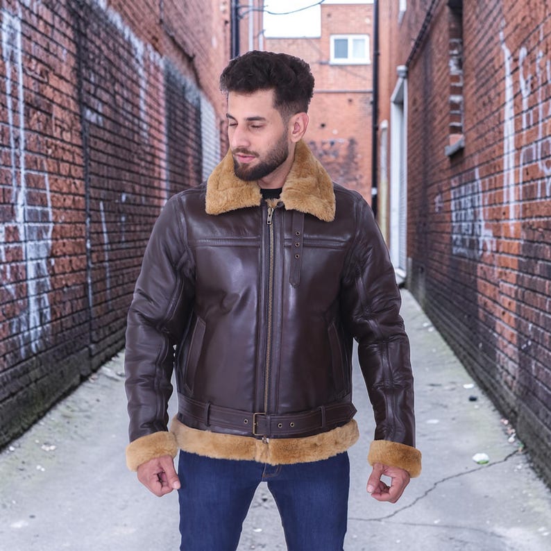 May include: A brown leather jacket with a tan shearling collar and cuffs. The jacket features a front zipper, a belt at the waist, and a classic aviator style. The model is wearing blue jeans.