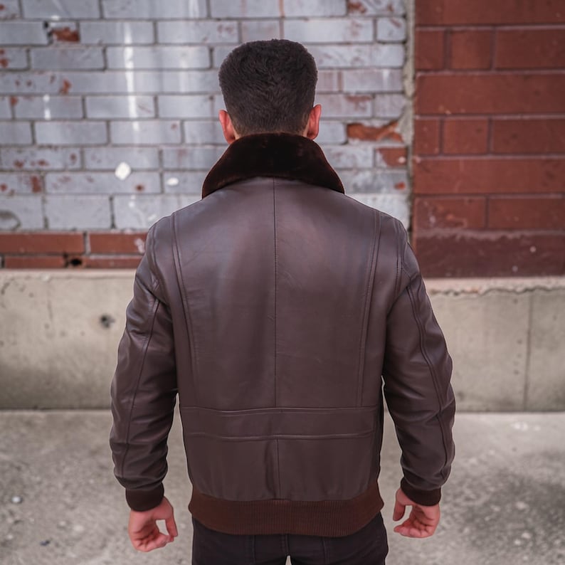 Puede incluir: Chaqueta bomber de cuero marr&oacute;n con cuello de piel marr&oacute;n oscuro. La chaqueta tiene dobladillo y pu&ntilde;os acanalados. La persona est&aacute; de espaldas, mostrando la parte trasera de la chaqueta. El fondo es una pared de ladrillos.