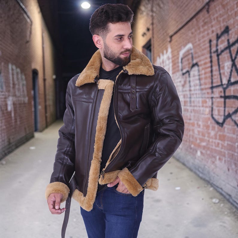 May include: A brown leather jacket with a tan shearling collar and lining. The jacket has a zipper closure, a buckled strap at the waist, and a classic aviator style. The jacket is worn with blue jeans.