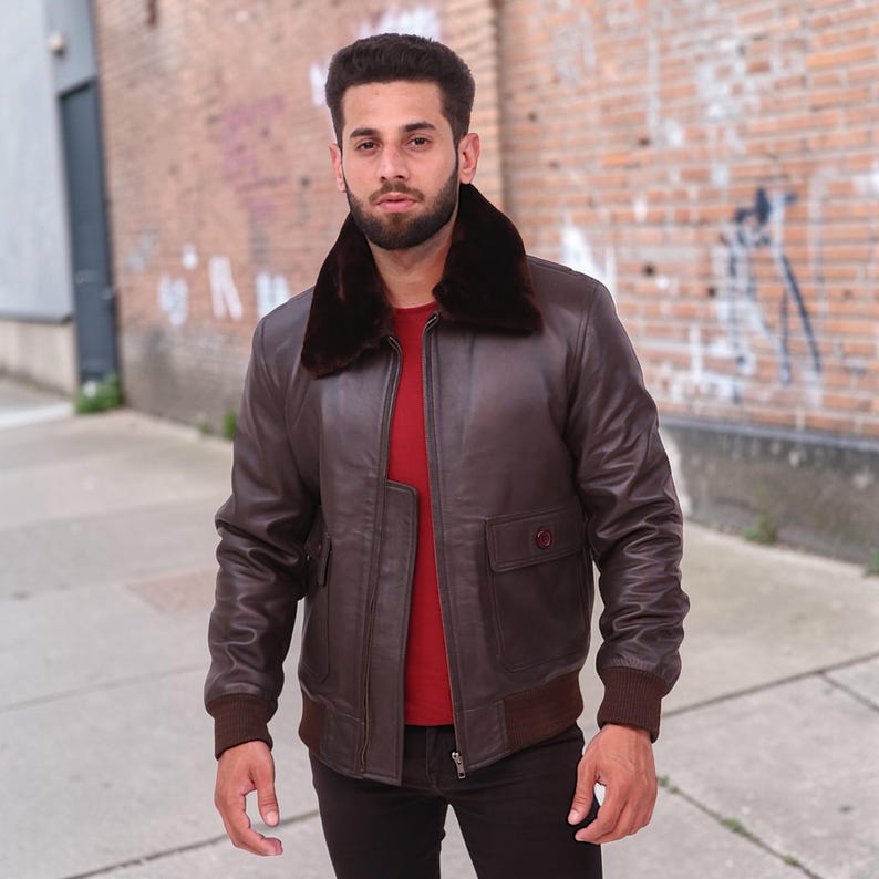 Puede incluir: Chaqueta bomber de cuero marr&oacute;n con cuello de piel sint&eacute;tica marr&oacute;n oscuro. La chaqueta tiene dos bolsillos delanteros, dobladillo y pu&ntilde;os acanalados. El modelo lleva una camisa roja debajo. La chaqueta est&aacute; cerrada.