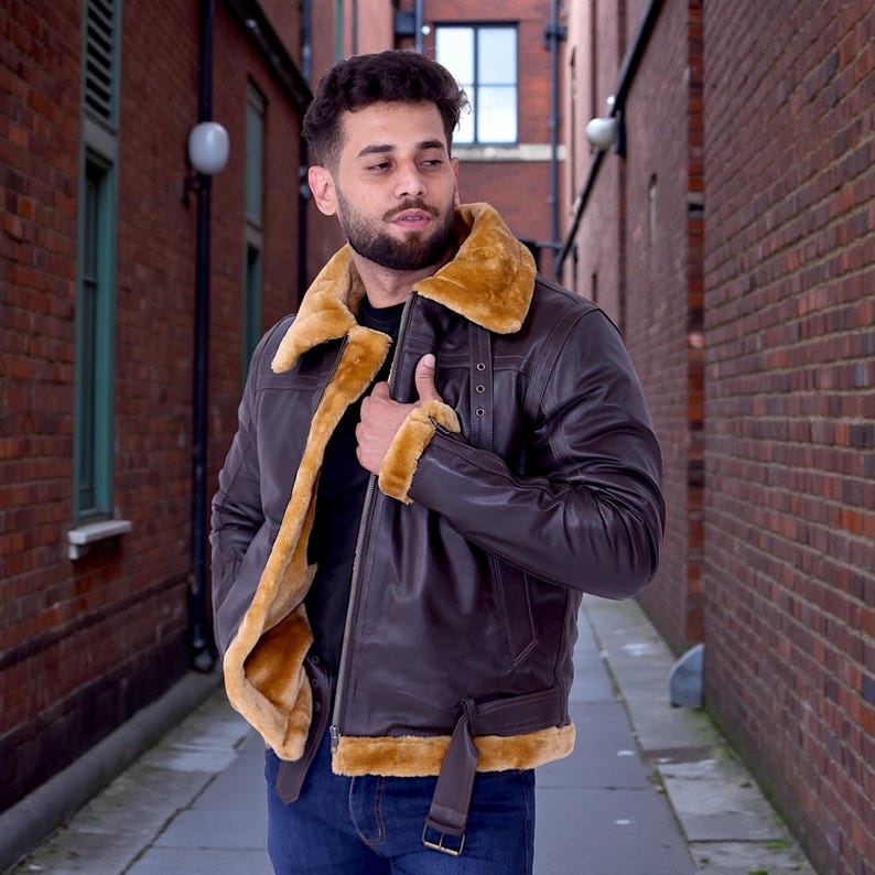 May include: A brown leather jacket with a tan shearling collar and lining. The jacket features a front zipper, a belt at the waist, and shearling trim on the cuffs. The jacket is worn over a black shirt and blue jeans.