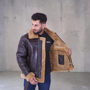 May include: A brown leather jacket with a tan shearling lining and trim. The jacket has a zip closure, a notched collar, and a buckle detail at the waist. The jacket is worn over a black shirt.