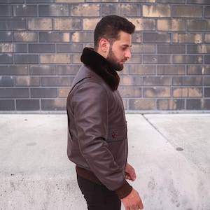 Puede incluir: Chaqueta bomber de cuero marr&oacute;n con cuello de piel marr&oacute;n oscuro. La chaqueta tiene dobladillo y pu&ntilde;os de canal&eacute; y un bolsillo abotonado. La chaqueta la lleva una persona de pie frente a una pared de ladrillos.