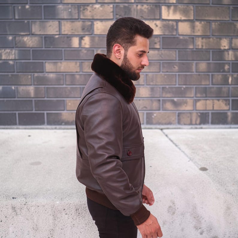 Puede incluir: Chaqueta bomber de cuero marr&oacute;n con cuello de piel marr&oacute;n oscuro. La chaqueta tiene dobladillo y pu&ntilde;os de canal&eacute; y un bolsillo abotonado. La chaqueta la lleva una persona de pie frente a una pared de ladrillos.
