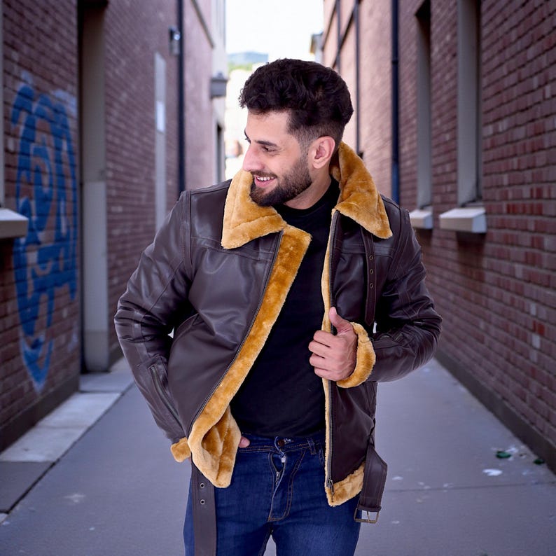 May include: A brown leather jacket with a tan shearling collar and lining. The jacket has a classic aviator style with a zip closure and a belt at the waist. The person is wearing a black shirt and blue jeans.