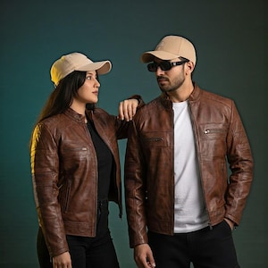 Puede incluir: Dos personas con chaquetas de cuero marrón y gorras de béisbol beiges. Las chaquetas tienen un estilo motero clásico con bolsillos con cremallera y detalles acanalados en las mangas. Una persona lleva gafas de sol. El fondo es un degradado de verde azulado y verde oscuro.