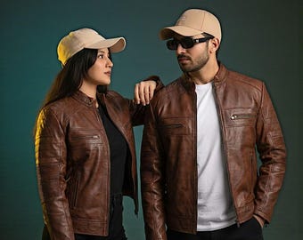 May include: Two people wearing brown leather jackets and tan baseball caps. The jackets have a classic biker style with zippered pockets and ribbed detailing on the sleeves. One person is wearing sunglasses. The background is a gradient of teal and dark green.