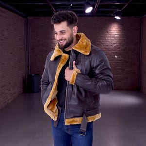 May include: A brown leather jacket with a tan shearling collar and lining. The jacket features a zip closure, a buckled belt at the waist, and shearling trim on the cuffs. The person is wearing blue jeans and a black shirt.