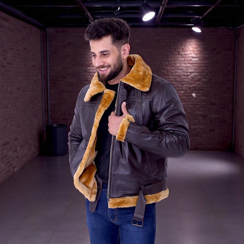 May include: A brown leather jacket with a tan shearling collar and lining. The jacket features a zip closure, a buckled belt at the waist, and shearling trim on the cuffs. The person is wearing blue jeans and a black shirt.