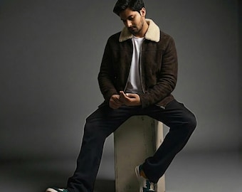 May include: A person wearing a brown suede jacket with a cream-colored shearling collar, a white t-shirt, and black pants. The person is wearing sneakers with green and yellow accents. The person is sitting on a white cube against a gray background.