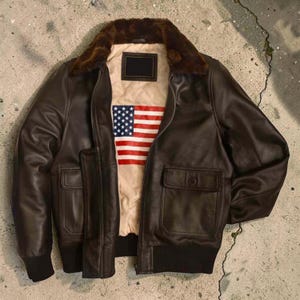 Puede incluir: Chaqueta bomber de cuero marrón con cuello de piel y dos bolsillos delanteros. El forro interior de la chaqueta presenta la bandera americana. La chaqueta tiene dobladillo y puños de canalé.