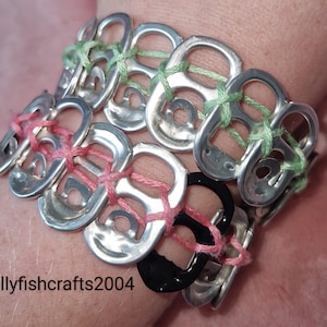 Puede incluir: Pulsera hecha a mano con anillas de latas de color plateado, unidas con hilo rosa y verde. La pulsera es un accesorio único, que muestra una reutilización creativa de materiales. El texto "jellyfishcrafts2004" es visible.