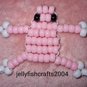 Puede incluir: Una figura rosa hecha a mano con cuentas, con brazos y piernas blancos y ojos negros. La figura está hecha de pequeñas cuentas redondas ensartadas con hilo blanco. El texto "jellyfishcrafts2004" está en la parte inferior.