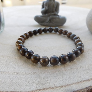 Peut inclure: Un bracelet de perles marron à l'aspect naturel et terreux. Le bracelet est composé de perles rondes et polies, probablement des pierres précieuses, et est présenté sur une surface en bois de couleur claire. Une petite statue de Bouddha assis est en arrière-plan.