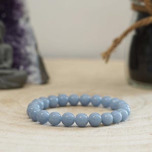 Peut inclure: Un bracelet de perles bleu clair. Le bracelet est composé de perles rondes et lisses. Le bracelet repose sur une surface en bois de couleur claire. Une petite statue et un pot sont en arrière-plan.
