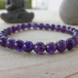 May include: A bracelet made of polished, round amethyst beads. The deep purple beads are strung together to form a flexible band. The bracelet is shown on a light-coloured, textured surface, with a blurred background.
