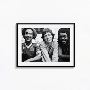 May include: A black and white framed photograph featuring three individuals smiling. The person on the left wears a knit cap and a collared shirt with a circular badge. The center person has shoulder-length hair, and the right person wears a beanie.