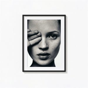 May include: A black and white framed wall art print featuring a close-up portrait of a face with one hand partially covering the eye. The image displays high-contrast lighting, detailed skin texture, and a focused gaze, all encased in a thin black frame.