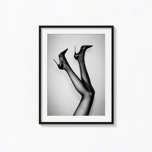 Op de afbeelding: Een zwart-wit ingelijste wandkunstprint met een paar benen in doorschijnende zwarte panty's en zwarte stilettohakken. De benen zijn naar boven gericht tegen een lichte achtergrond en bevinden zich in een dunne zwarte lijst aan de muur.