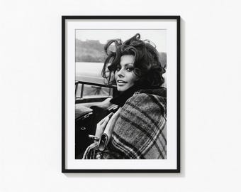 May include: A black and white framed photograph featuring a person with voluminous dark hair wearing a plaid coat, looking over their shoulder while standing next to a car. The portrait is encased in a thin black frame with a white mat border.