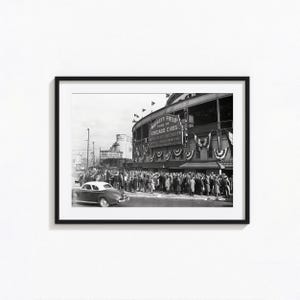 Può includere: Una fotografia incorniciata in bianco e nero del Wrigley Field, casa dei Chicago Cubs. Una grande folla si raduna davanti all'ingresso dello stadio, decorato con festoni. Un'auto d'epoca è parcheggiata in primo piano vicino al muro dello stadio.