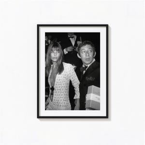 May include: A black and white framed photograph featuring a person with long hair wearing a white lace dress with a deep neckline, standing beside another person in a dark suit and bow tie, who is holding a rectangular object with a visible label.