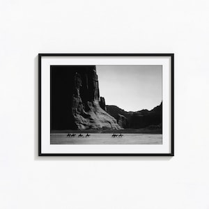 May include: A black and white framed wall art print featuring a landscape scene with a large rock formation in the background and a group of people riding horses across a flat, open desert terrain in the foreground, enclosed in a black frame.