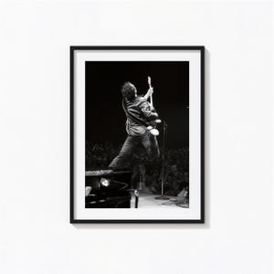 May include: A black and white framed photograph depicting a musician performing on stage, captured from behind while jumping and playing an electric guitar. The performer is silhouetted against a dark background with a crowd visible below.
