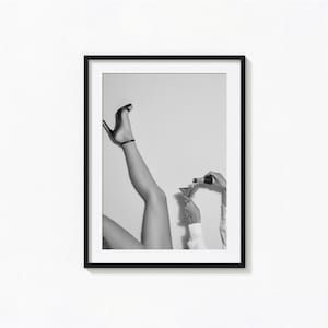 May include: A black and white framed wall art print featuring a pair of legs raised in the air while wearing a high-heeled shoe, alongside a hand pouring a drink from a bottle into a martini glass, presented in a simple black frame.