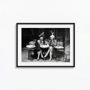 May include: A black and white framed photograph depicting two individuals seated at small round cafe tables outdoors. One person wears a cloche hat and dark coat, while the other wears a turban-style hat and coat. The word TELEPHONE is visible.