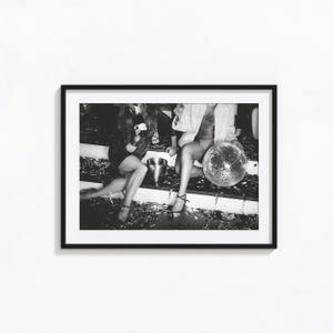 May include: A black and white framed photograph depicting two individuals sitting on stairs covered in confetti. One person holds a bottle, while the other holds a reflective disco ball. The scene captures a festive atmosphere with scattered debris.