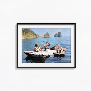 May include: A framed photographic print showing three individuals relaxing on brown inflatable rafts in blue ocean water. In the background, large rocky cliffs rise from the sea under a clear sky. The scene captures a leisure moment on the water.