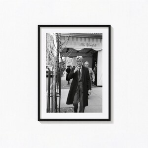 Op de afbeelding: Een zwart-wit ingelijste foto van een persoon in een lange donkere jas en broek die op een trottoir voor hotel The Carlyle loopt. De persoon houdt een sigaret bij het gezicht, met andere figuren zichtbaar op de achtergrond.