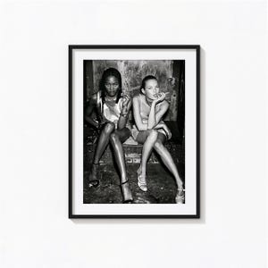 May include: A black and white framed photographic print featuring two individuals sitting side by side. Both are wearing sleeveless tops and light-colored footwear. One individual holds a cigarette, while the other holds a cigarette to their lips. The scene is captured in a high-contrast, monochromatic style within a simple black frame.