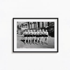 May include: A black and white framed photograph featuring seven individuals standing in a row, each wearing a white shirt with a single black letter spelling out NEWYORK. They are dressed in dark shorts and bow ties, positioned in front of a building.