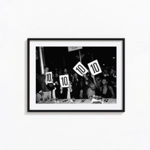 Könnte beinhalten: Ein schwarz-weiß gerahmtes Foto, das eine Gruppe von Menschen zeigt, die weiße Schilder mit der Zahl 10 in fetter schwarzer Schrift hochhalten. Die Szene zeigt eine Jury-Veranstaltung, bei der Personen an einem Tisch sitzen und ihre Punktzahlen einem Publikum in einer dunklen Umgebung präsentieren.