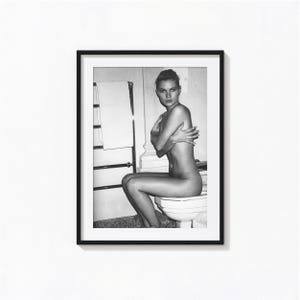 May include: A black and white framed photographic print featuring a person sitting on a toilet in a bathroom setting. The person is positioned in profile, facing the camera with arms crossed over their chest. A towel rack is visible in the background.