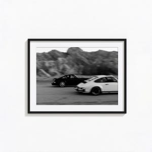 May include: A black framed wall art print featuring a black and white motion-blurred photograph of two sports cars driving on a road with a rocky landscape in the background. The image captures a dark car and a light car in side-by-side motion.