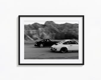 Op de afbeelding: Een ingelijste wandkunstprint in zwart met een zwart-witfoto met bewegingsonscherpte van twee sportwagens op een weg met een rotsachtig landschap op de achtergrond. De afbeelding toont een donkere auto en een lichte auto in zijwaartse beweging.