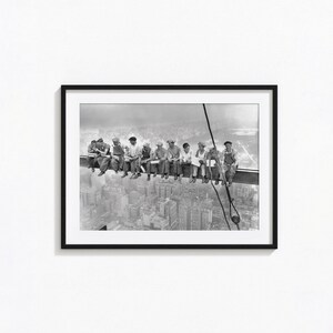May include: A black and white framed photograph depicting eleven construction workers sitting on a steel beam high above a city skyline. The workers are dressed in work clothes and caps, eating lunch while suspended over the urban landscape below.