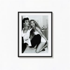 May include: A black and white framed photograph featuring two individuals in a bathroom setting. One individual wears an unbuttoned white shirt and dark trousers, while the other wears a white tank top and shorts, positioned near a tiled wall.