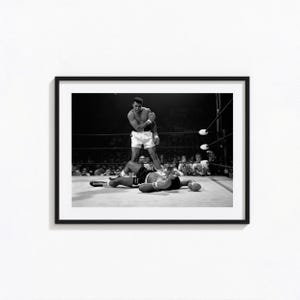 Könnte beinhalten: Ein schwarz-weiß gerahmtes Foto, das einen Boxkampf zeigt. Ein stehender Boxer in weißen Boxshorts und Handschuhen steht über einem Gegner, der auf dem Boden liegt. Die Szene spielt in einem Boxring mit Seilen, umgeben von Zuschauern.