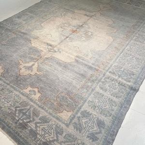 以下が含まれることがあります： 色あせたヴィンテージ感のある、長方形の大きなラグ。ラグは、クリームとピーチの色調の中央のデザインが特徴で、くすんだ青色のフィールドと幾何学模様の細部が施されたボーダーで囲まれています。 全体的な美しさは柔らかく、魅力的です。