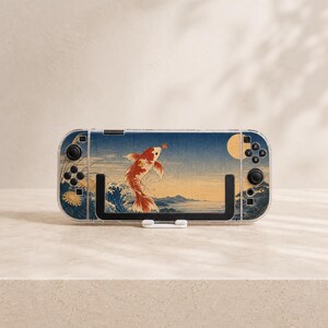 May include: A handheld gaming console featuring a clear protective case and a decorative skin. The skin displays a Japanese-inspired art print of a red and white koi fish leaping from blue waves toward a yellow moon against a dark blue sky.